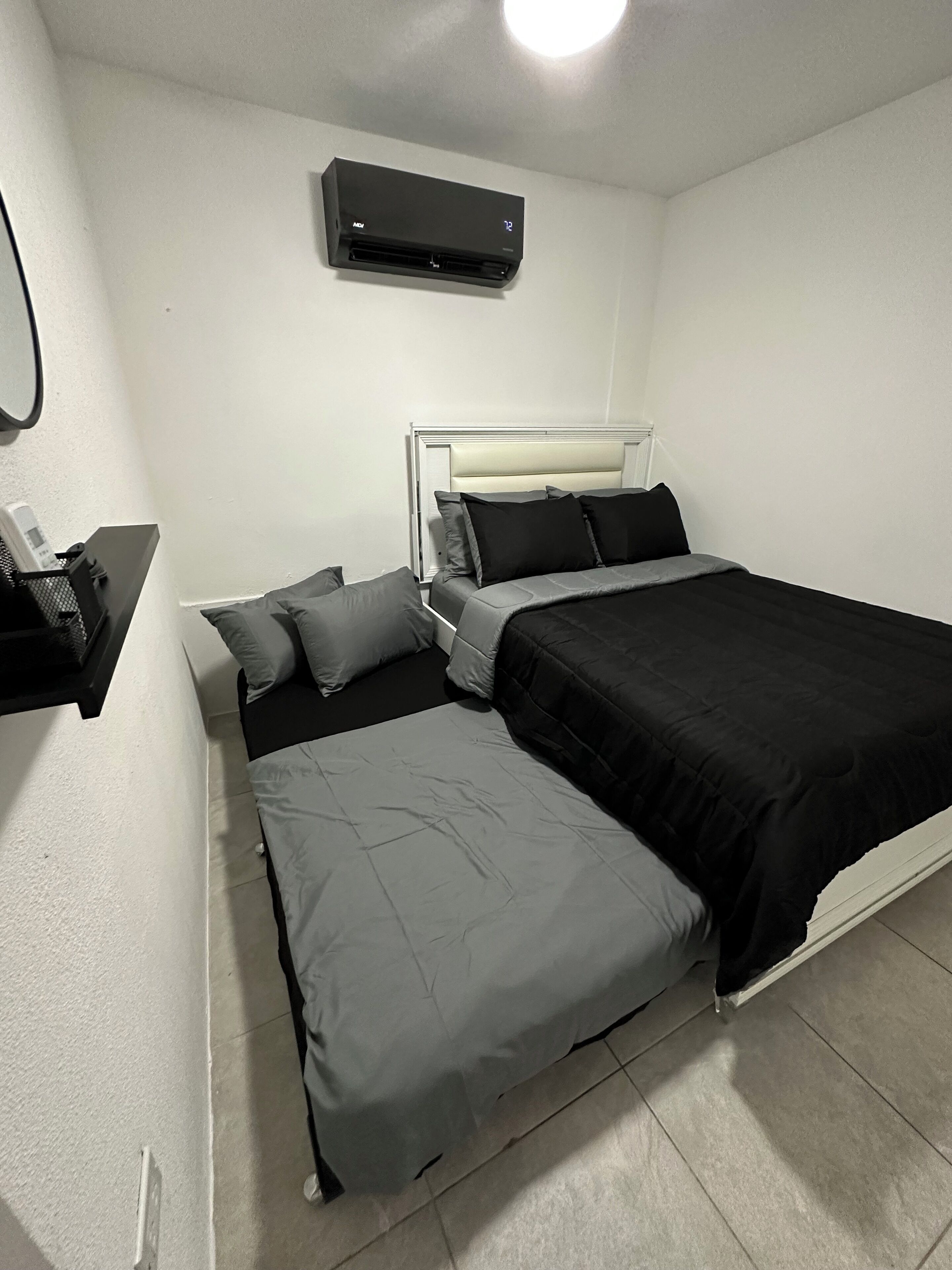 A bedroom with a bed, pillows, a bedside table, a wall-mounted air conditioner, and a small shelf.