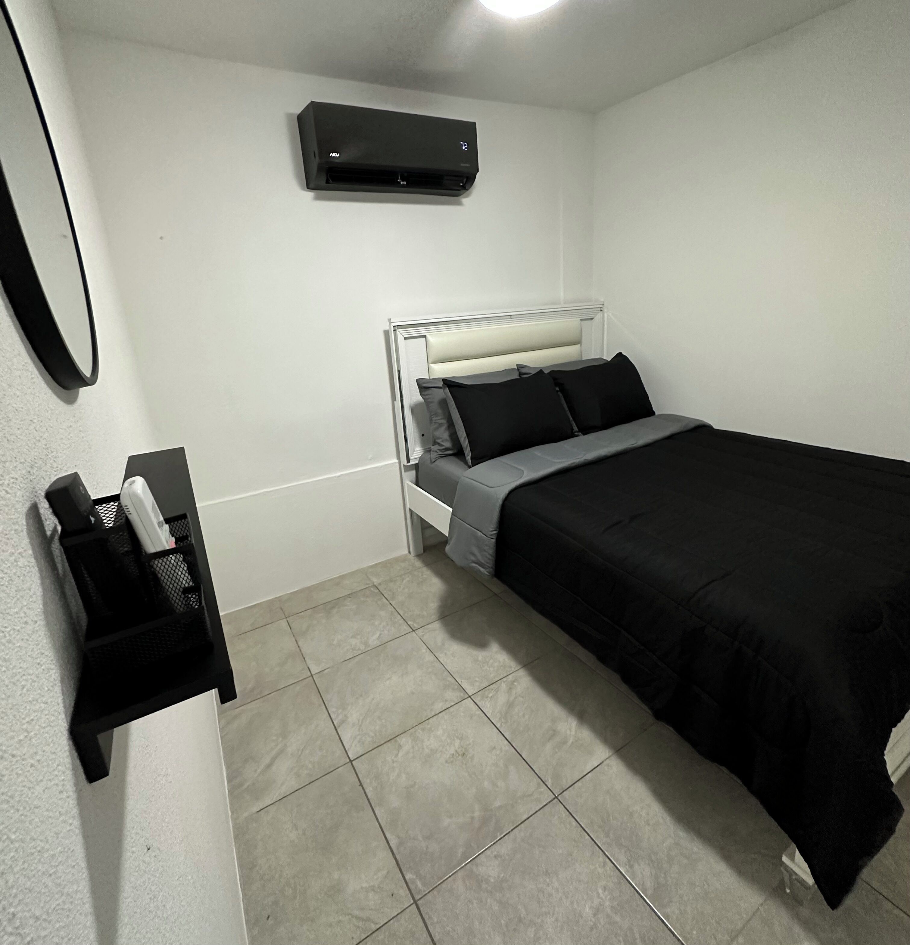 A small, clean room with a bed, a bedside table, a wall-mounted air conditioner, and a black storage unit with a mirror.