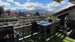En balkon med et bord og stole, et overdækket område og udsigt til bygninger og bjerge.