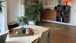 Spiseområde med et bord, stole, hængende lyspærer, en potteplante og en reol med bøger og dekorative genstande.