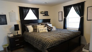 A bedroom with a large bed, dark curtains, a bedside table with a lamp, and a small trash bin. - Whole House in N. Denver with hot-tub (Denver)
