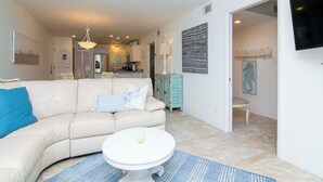 Un salon moderne avec un canapé beige, une table basse ronde blanche et un tapis bleu. On aperçoit une cuisine à l’arrière-plan, avec des meubles blancs et un coin repas.