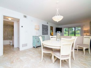 A dining area with a round table and chairs, a chandelier, and a view of the outdoors through large windows.