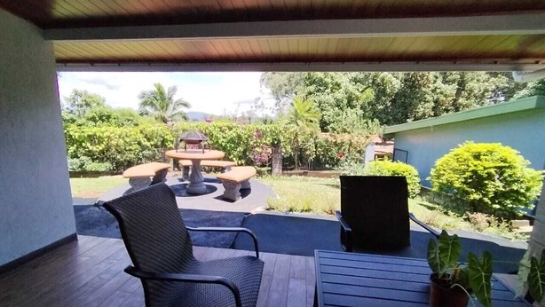 A covered patio with a table and chairs, a potted plant, and a view of a garden with a table and chairs in the background.