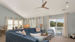 A spacious living room with a large blue sectional sofa, a ceiling fan, and a view to the outside. - SemiSecluded OBX Beach House, Heated Pool&Hot Tub, Beach Access, Pinball, Arcade (Corolla)