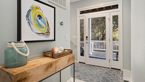 A hallway with a wooden console table, a framed abstract painting, a basket with a rope handle, and a door leading to an outdoor area with a railing. - SemiSecluded OBX Beach House, Heated Pool&Hot Tub, Beach Access, Pinball, Arcade (Corolla)
