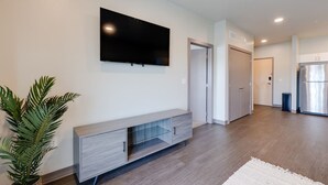 A modern living room with a flat-screen TV mounted on the wall, a grey entertainment unit, and a potted plant. - Cozy 1B1B w/Wi-Fi, Balcony Views, Coffee + Parking (Des Moines)