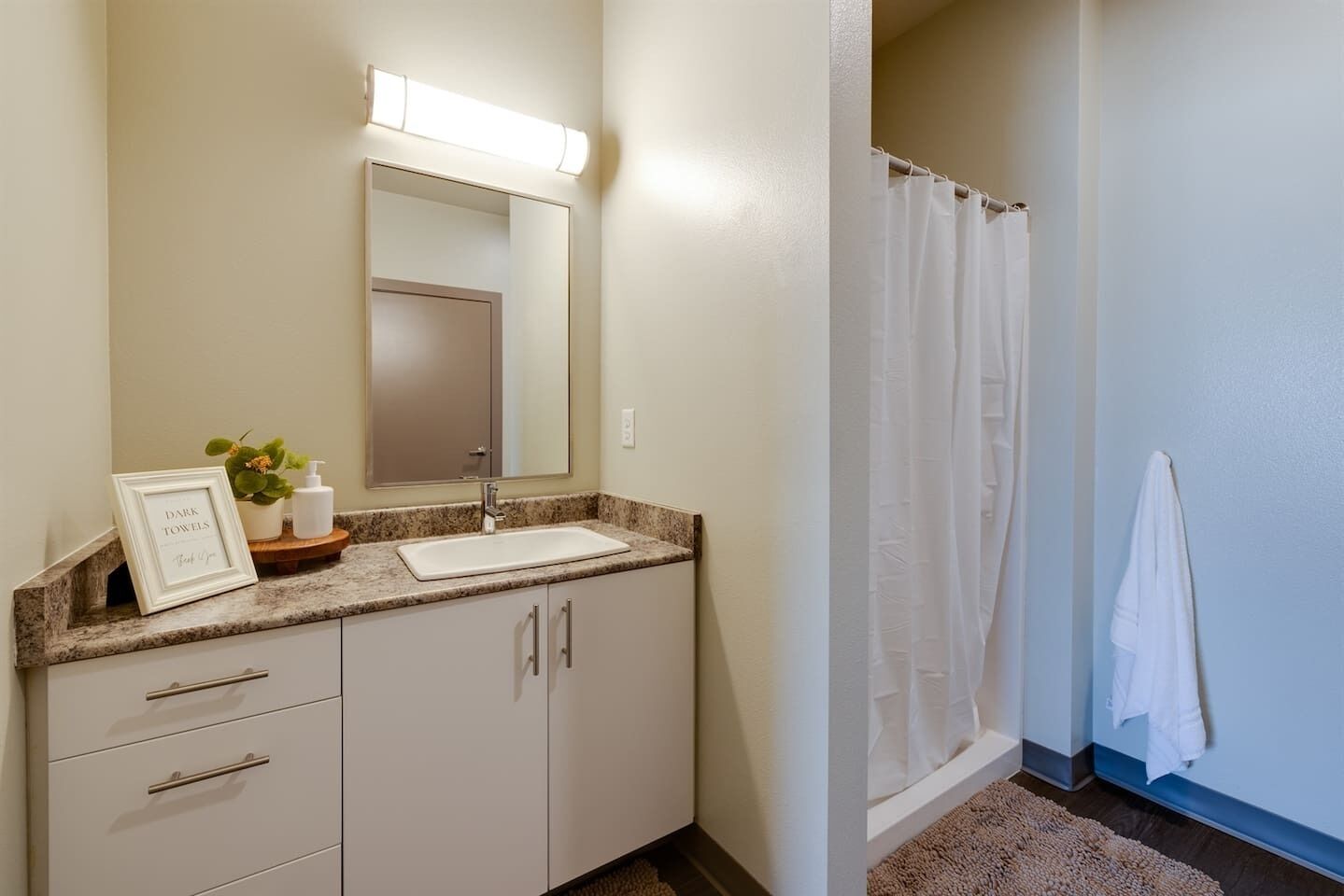 A bathroom with a sink, mirror, and shower curtain. There is a towel hanging on the wall and a small plant on the counter.