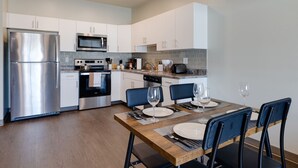 A modern kitchen with stainless steel appliances, white cabinetry, and a dining area with a wooden table and blue chairs. - Cozy 1B1B w/Wi-Fi, Balcony Views, Coffee + Parking (Des Moines)