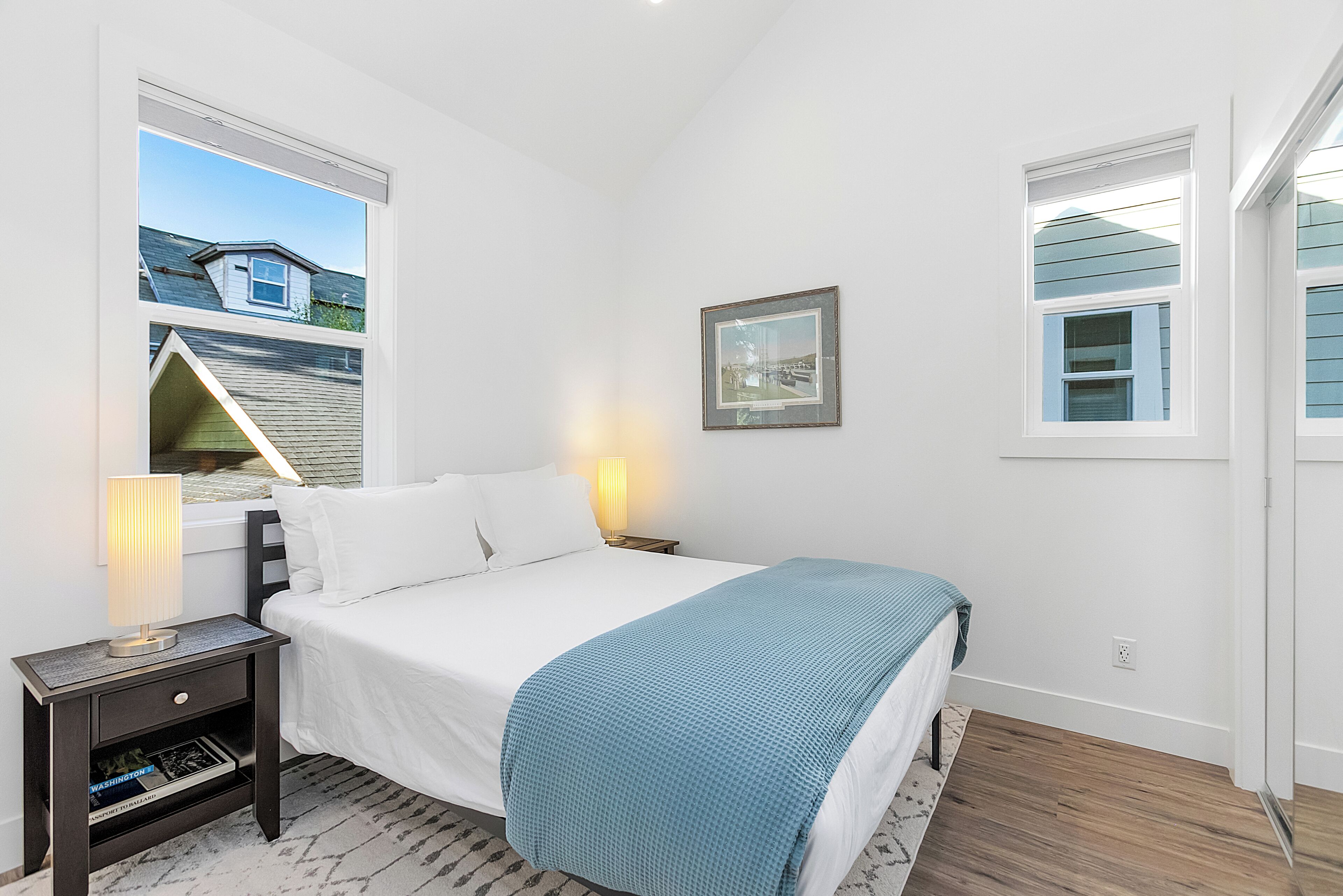 A bedroom with a bed, nightstand, lamp, framed picture, and a window with a view of a house.