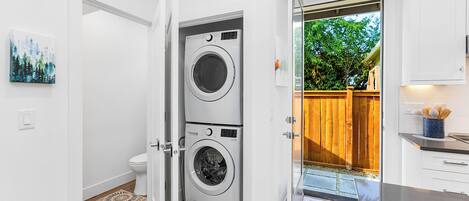 A laundry room with two stacked front-loading washing machines, a white toilet, and a wooden door leading to an outdoor area with a wooden fence.