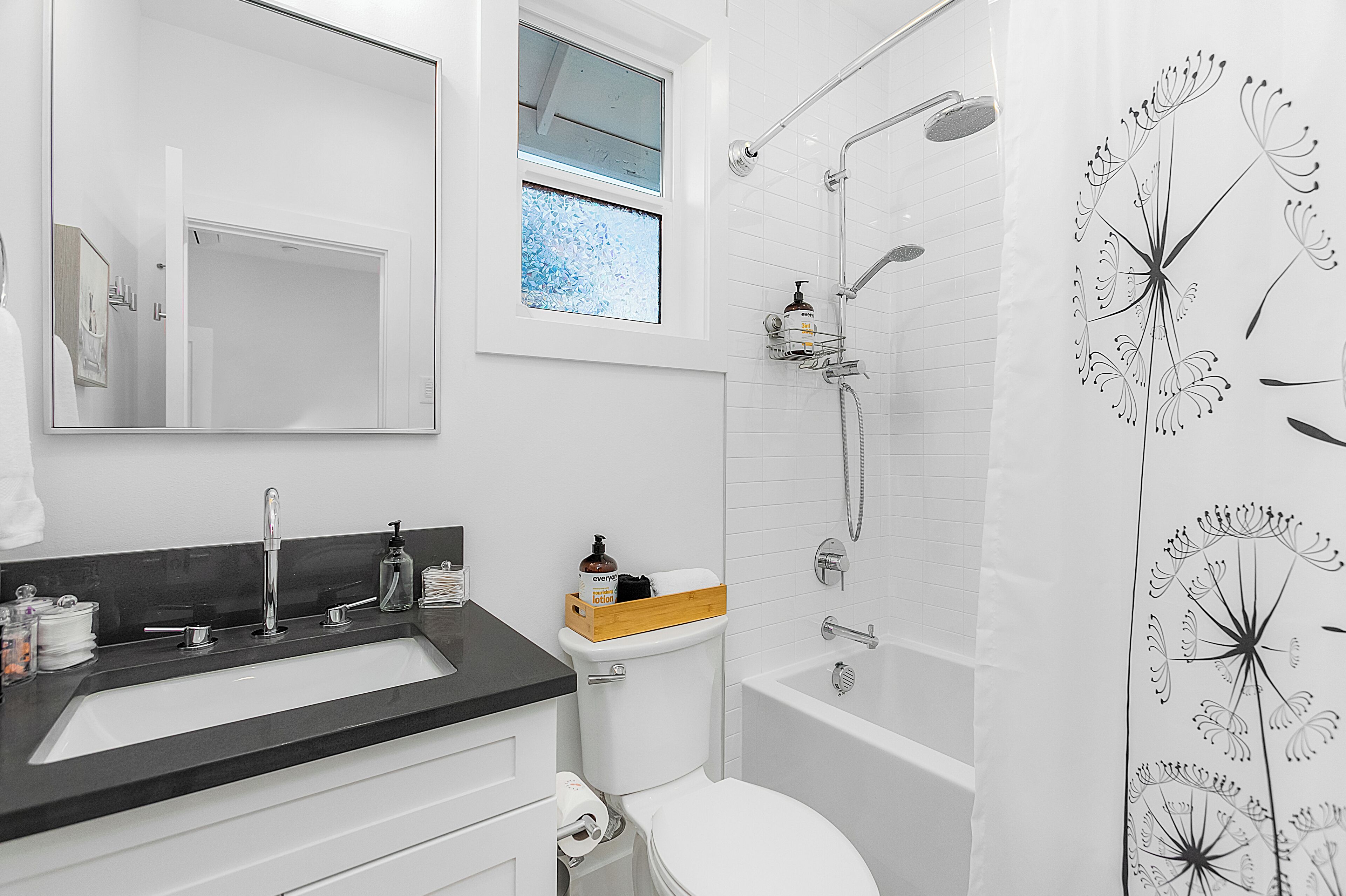 A modern bathroom with a large mirror, a black countertop sink, a toilet, a shower with a rain showerhead, and a window with frosted glass.