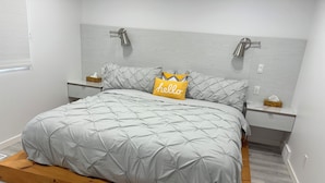 Un dormitorio moderno con cama sobre plataforma de madera, ropa de cama gris, dos mesitas de noche con lámparas y una pared blanca con interruptor de luz.