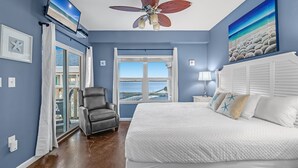 A bedroom with a large bed, a ceiling fan, a TV, and a view of the beach through a window.