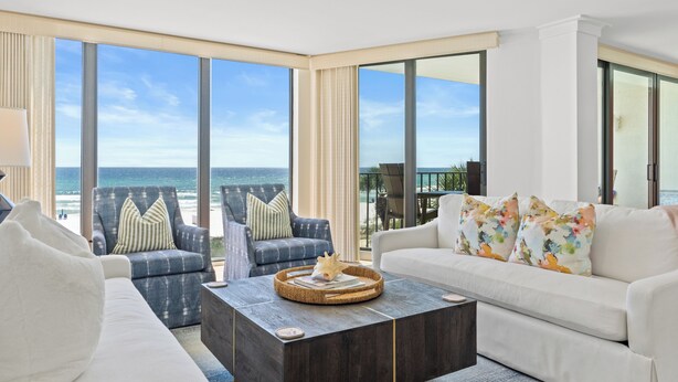 A modern living room with a large window overlooking the ocean, a white sofa, a blue armchair, and a coffee table.