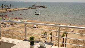A balcony with a table and chairs overlooking a beach with umbrellas, palm trees, and a pier. - Wave Beachfront Apartments (Durrës)