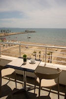 Balcón con mesa y sillas con vista a una playa con sombrillas, palmeras y un muelle.