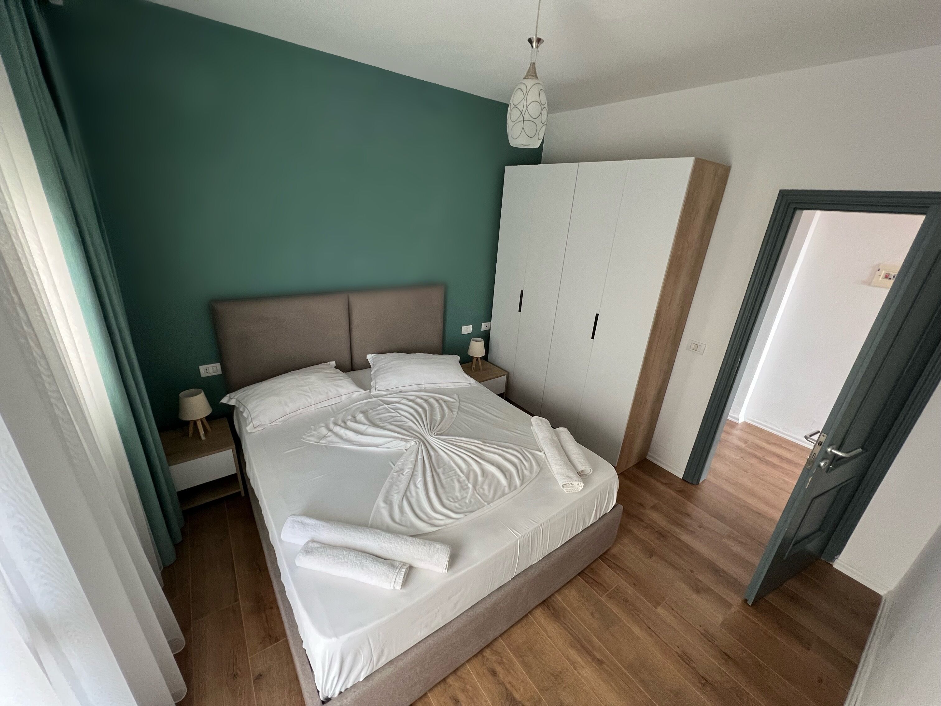 A neatly made bed with white linens and towels, a wooden headboard, a white wardrobe, and a wooden floor.
