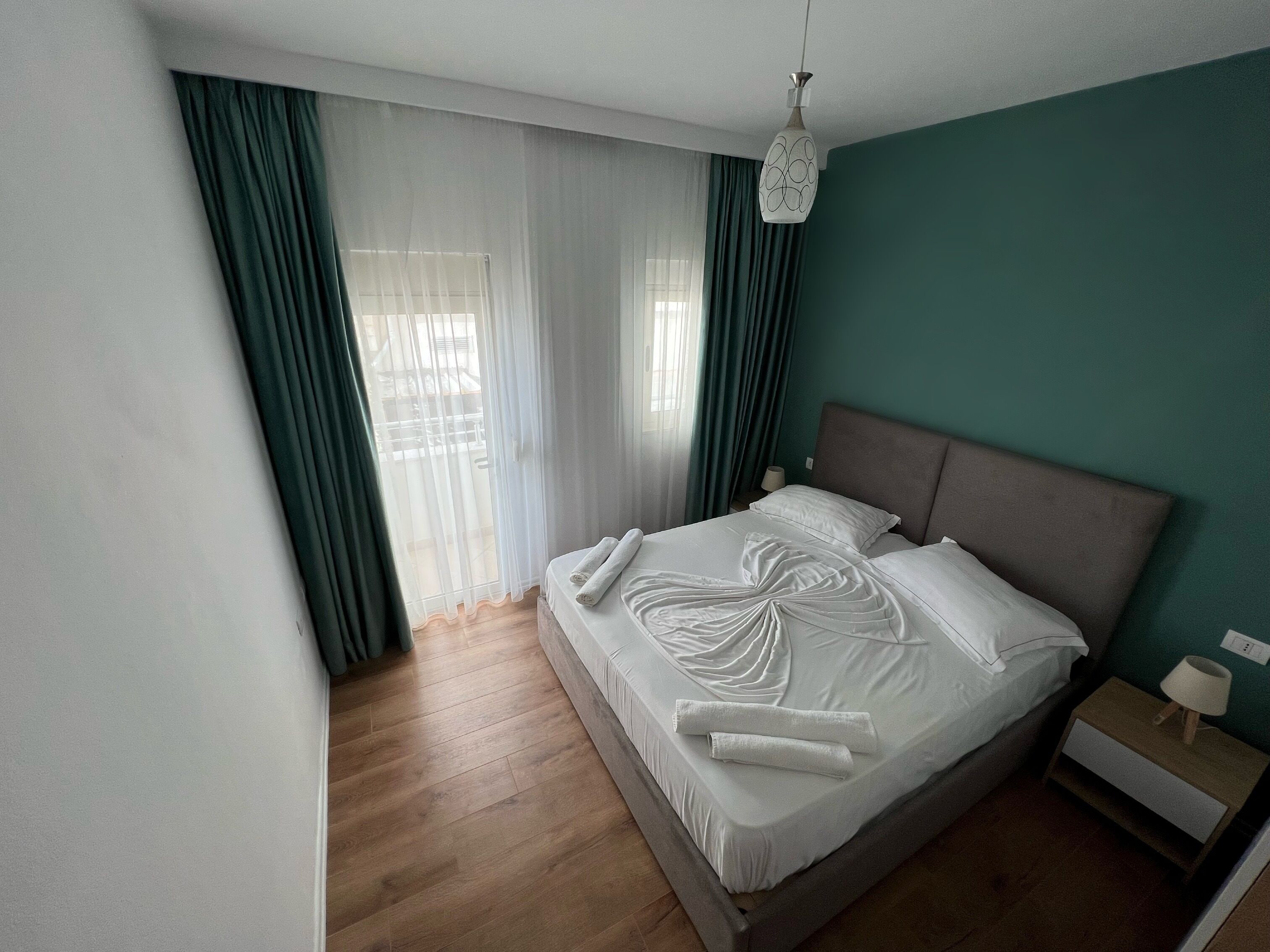 A modern hotel room with a large bed, a bedside table with a lamp, and sheer curtains allowing natural light.