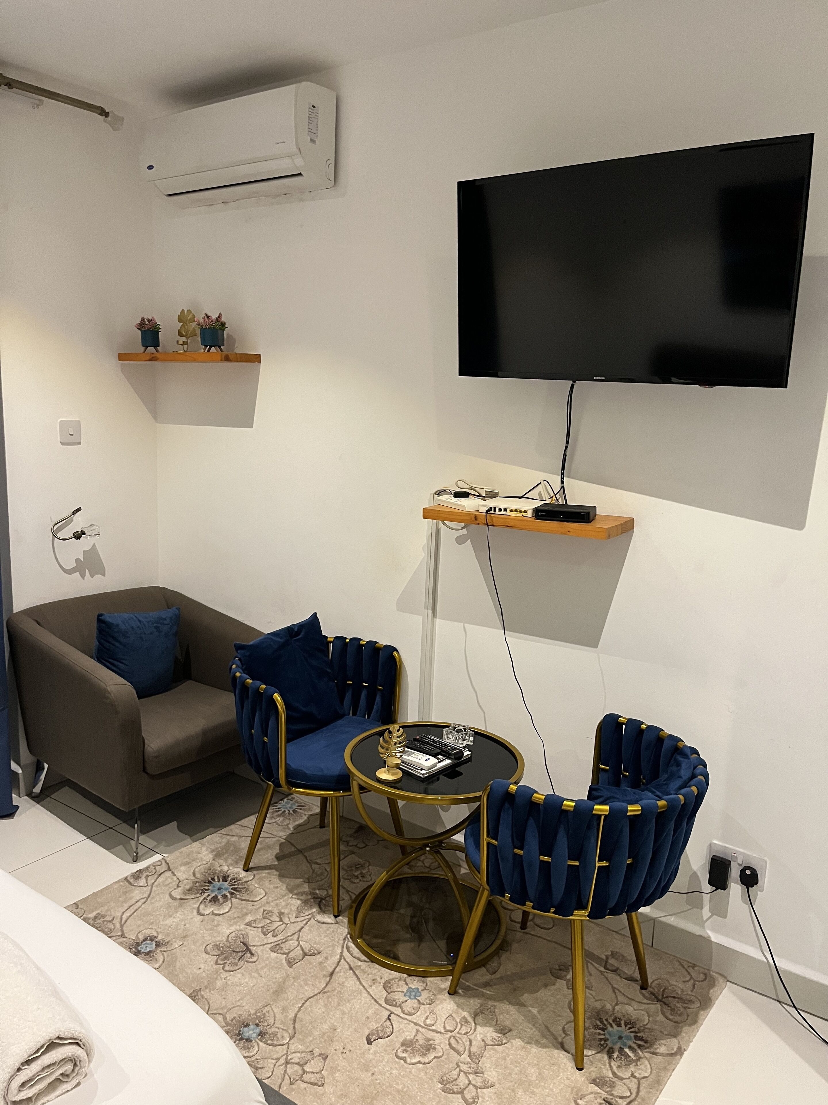 Una sala de estar moderna con un televisor de pantalla plana montado en la pared, un sofá, dos sillones azules, una pequeña mesa de centro y una estantería decorativa con plantas.