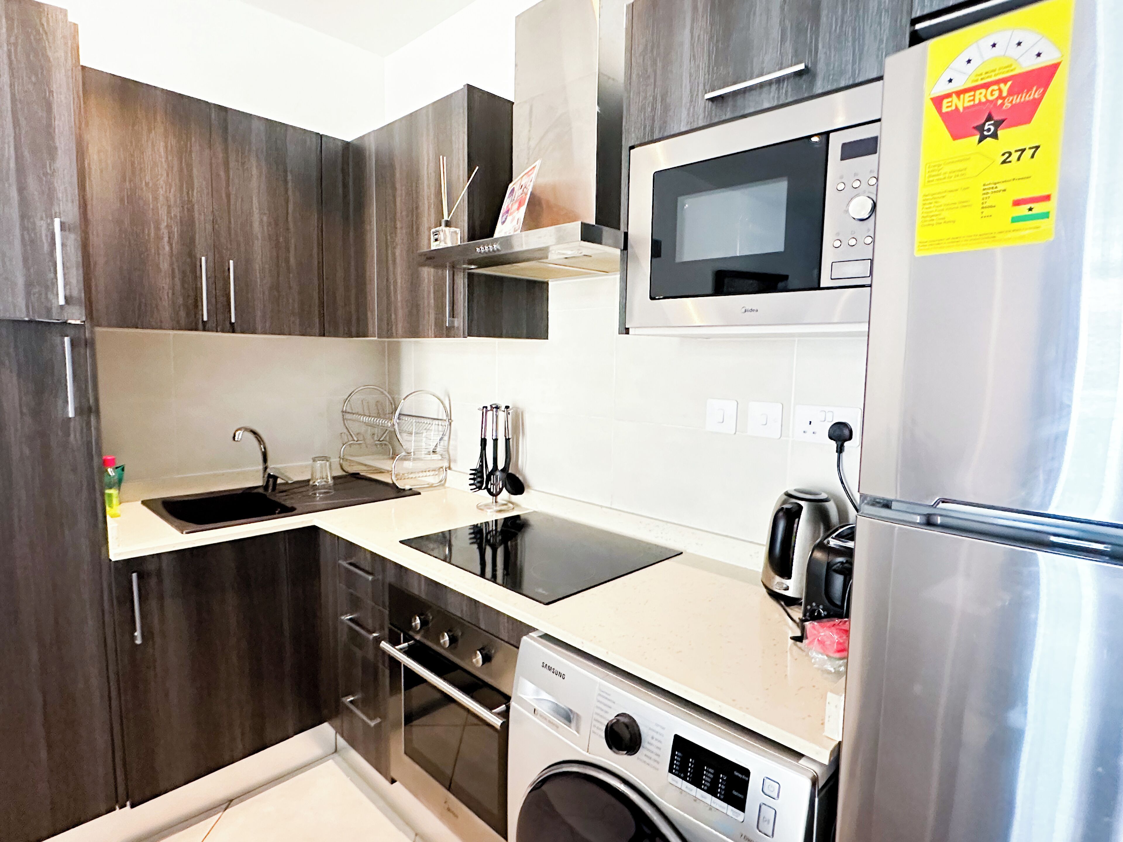 A compact kitchen with dark wood cabinets, a stainless steel refrigerator, a microwave, a built-in oven, and a washing machine.