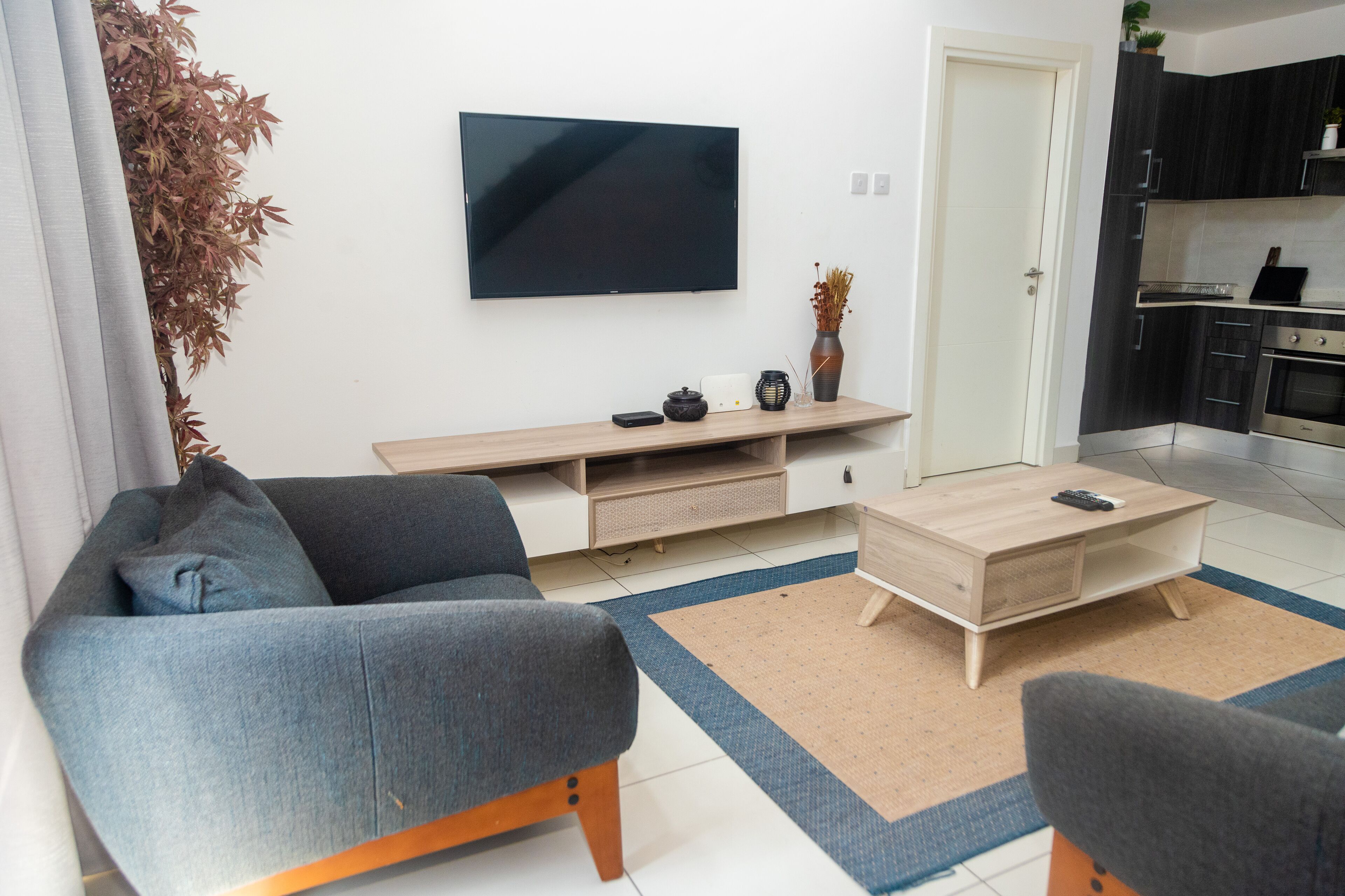 A modern living room with a flat-screen TV, a sofa, a coffee table, and a kitchen area in the background.