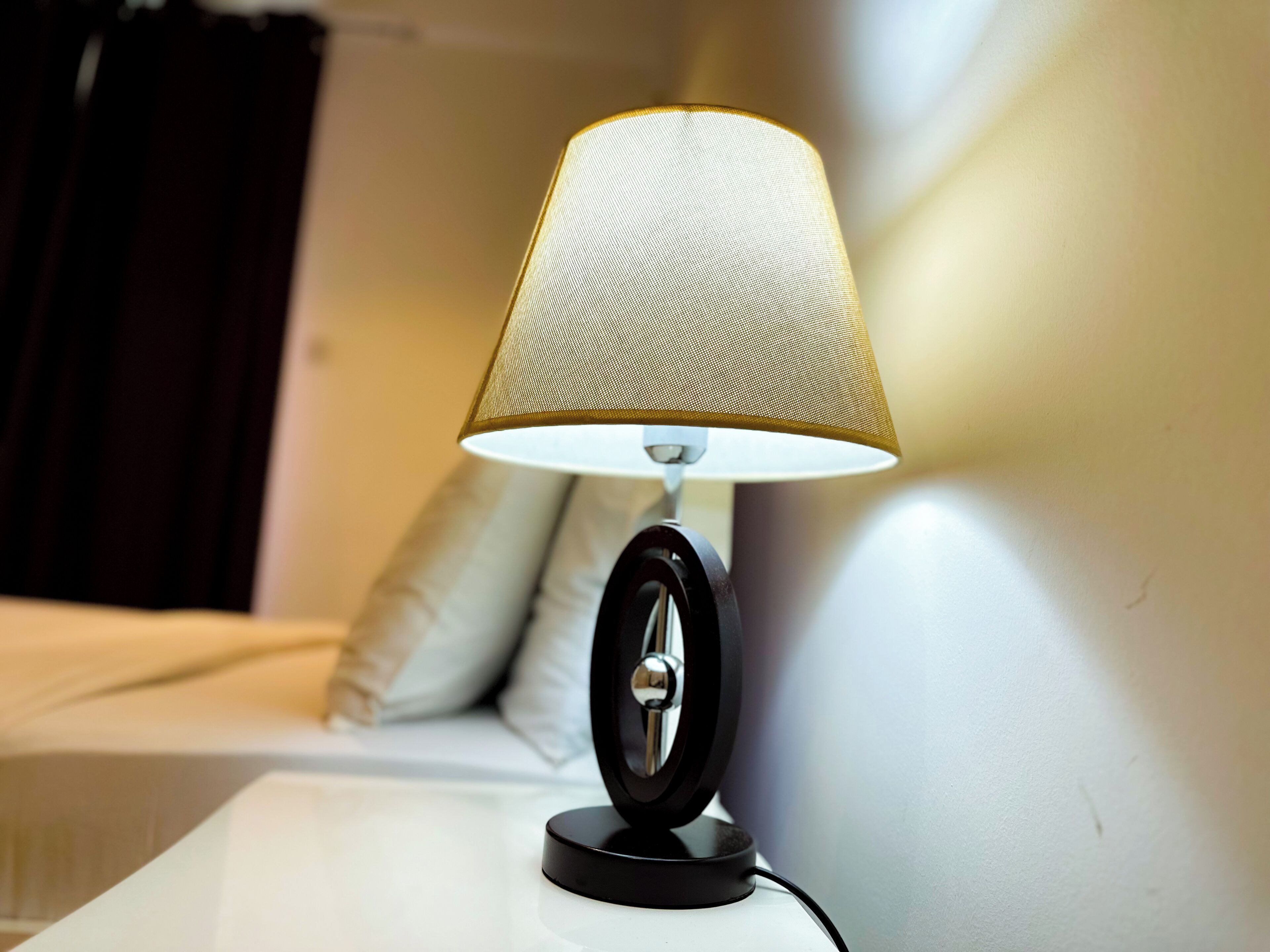 Uma mesa de cabeceira com uma luminária, uma cama e uma almofada ao fundo.