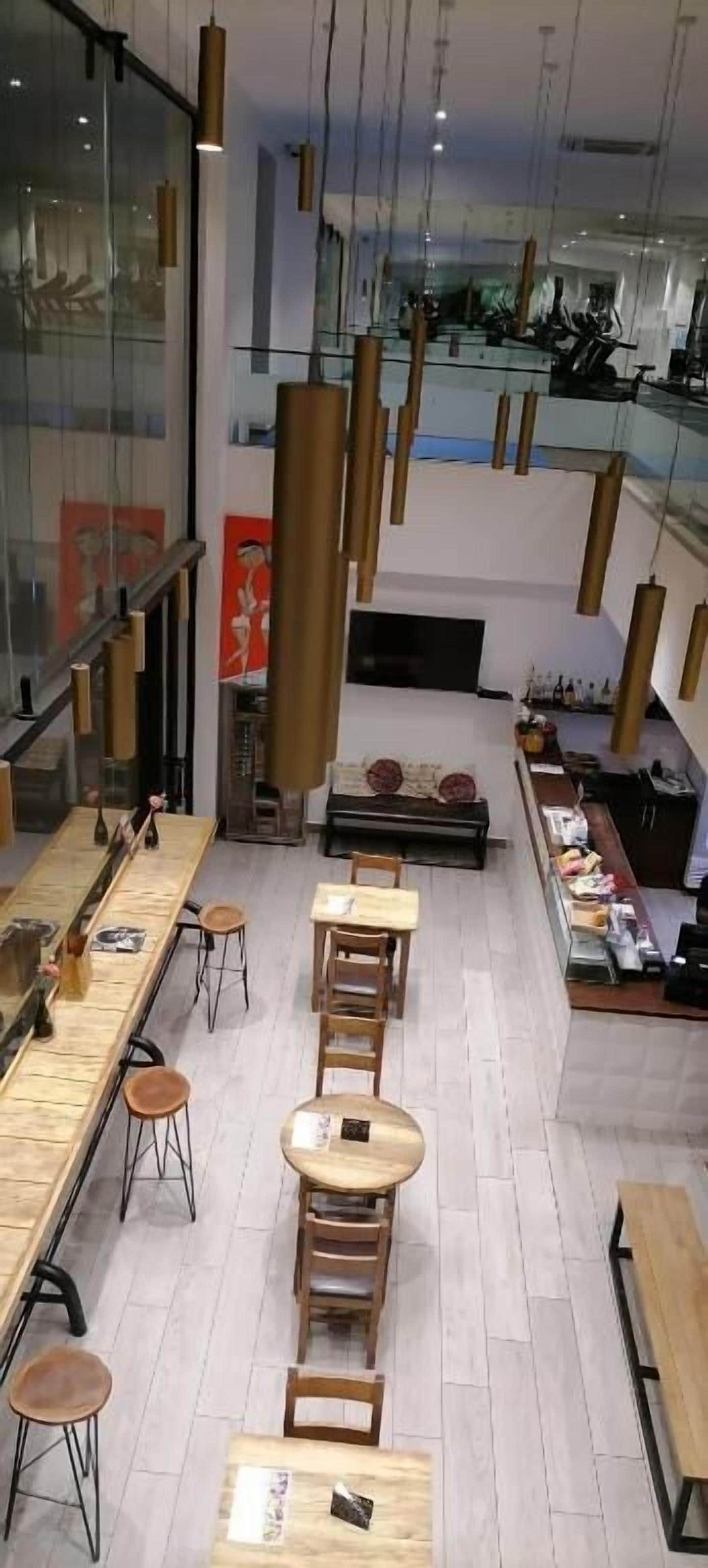 A modern café with wooden tables and chairs, hanging pendant lights, and a glass wall with artwork.