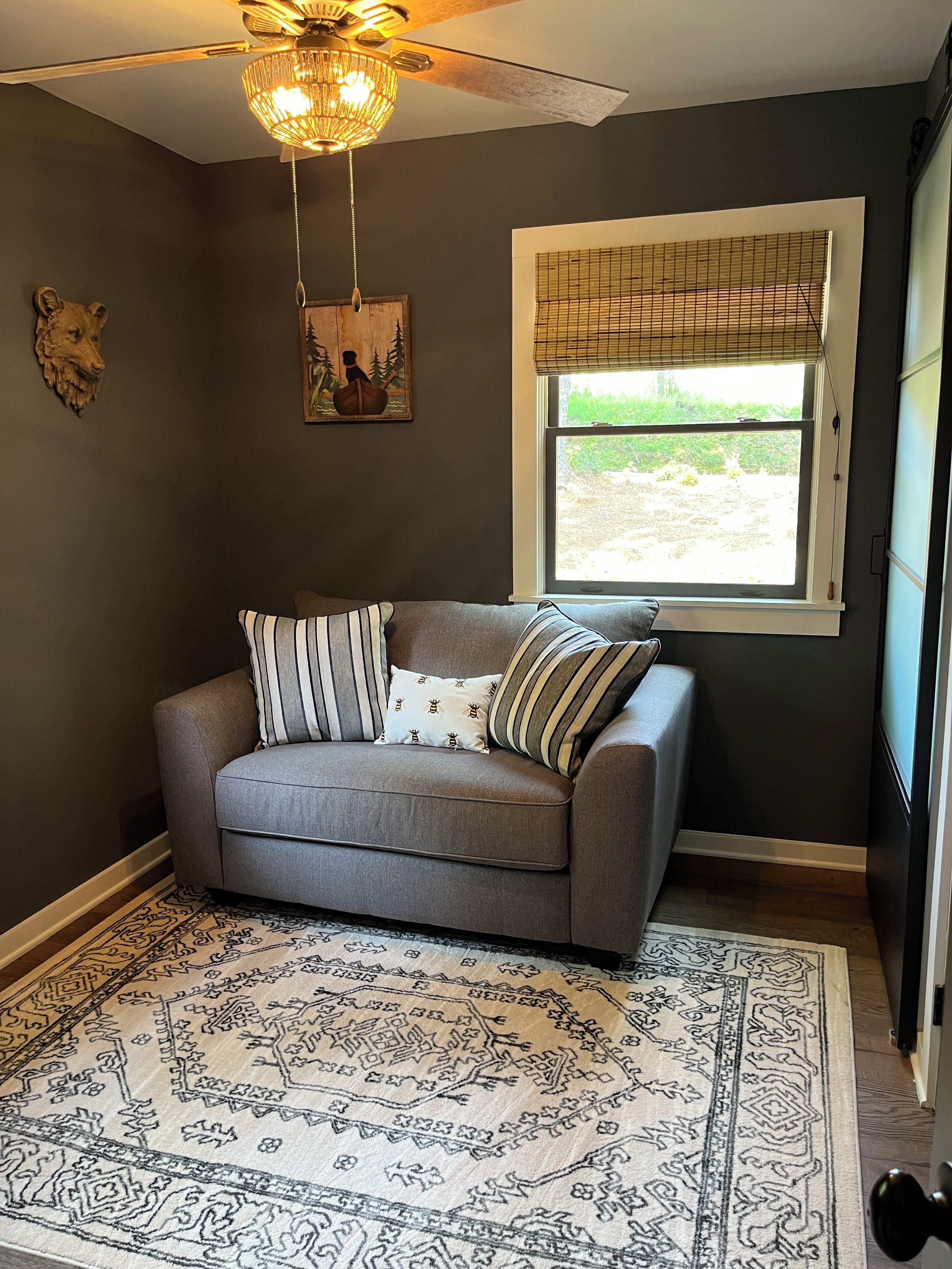 A room with a grey sofa, patterned rug, a ceiling fan, a window with blinds, and a wall-mounted artwork.