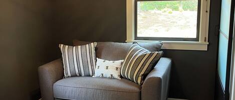 A room with a grey sofa, patterned rug, a ceiling fan, a window with blinds, and a wall-mounted artwork.