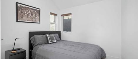 Quarto com cama, mesa de cabeceira, fotografia emoldurada e duas janelas.
