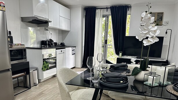 Et moderne køkken med hvide skabe, et spiseområde med et glasbord, og en stue med et fjernsyn og en plante.
