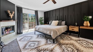 A modern bedroom with a fireplace, a bed with a headboard, a ceiling fan, and a large window. - Cozy Cabin Nestled in the Wilderness - 1 mile from Bear mountain (Big Bear Lake)