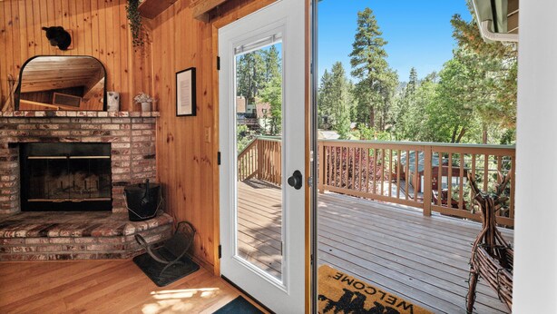 A wooden interior with a fireplace, a door leading to an outdoor deck, and a view of a forested area. - Cozy Cabin Nestled in the Wilderness - 1 mile from Bear mountain (Big Bear Lake)