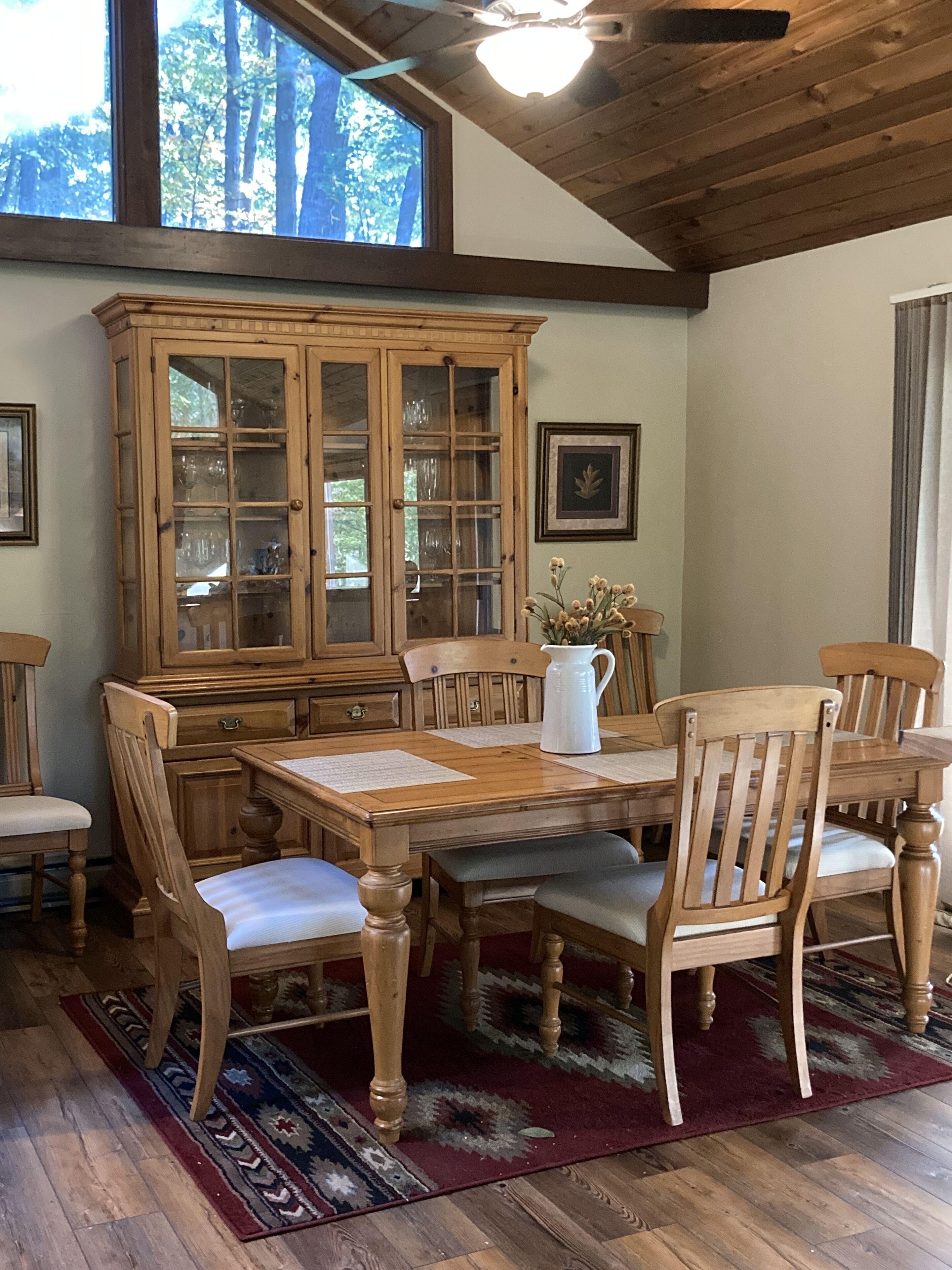 A dining area with a wooden table and chairs, a cabinet with glass doors, and a window with a view of trees.