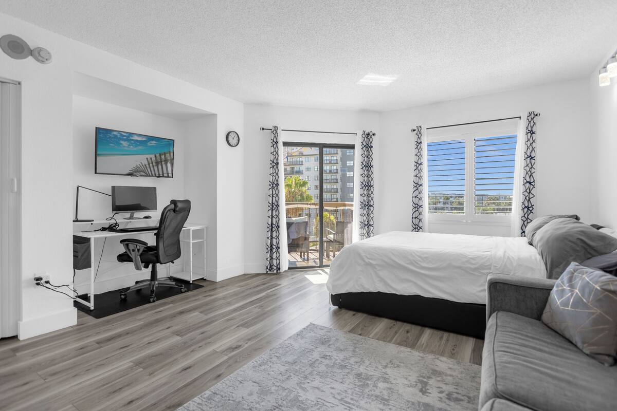 A modern bedroom with a bed, a desk with a computer, and a view of the outside balcony.