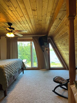 A bedroom with a bed, a ceiling fan, a TV mounted on the wall, and a view of the outdoors through a window.