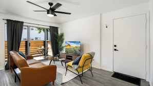 Un salon moderne avec un canapé, un fauteuil et un téléviseur posé sur un meuble. On y trouve également un ventilateur de plafond, une porte et une fenêtre avec des rideaux.