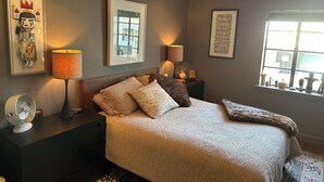 Une chambre à coucher comprenant un lit, deux tables de chevet, un ventilateur fixé au mur et une fenêtre avec des stores.
