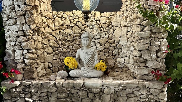 Une statue de Bouddha en pierre, surmontée d’un luminaire, et entourée de fleurs et de plantes.