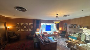 Un salon spacieux avec un grand canapé, des fauteuils et une table basse. On y trouve une cheminée avec un manteau de cheminée, ainsi qu’une grande fenêtre offrant une vue sur la ville.