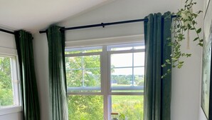 Een slaapkamer met een houten dressoir, groene gordijnen, een raam met uitzicht op groen en een hangplant.