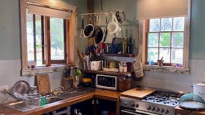 A compact kitchen with a gas stove, microwave, and a sink. There are hanging pots and pans, and a window with a view of greenery outside. - Little House in the Nursery (Wallabadah)