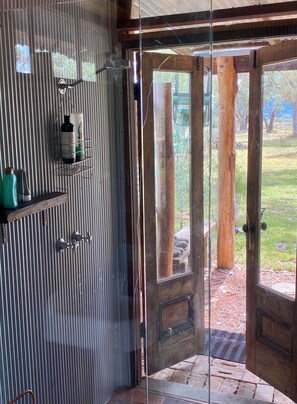 A room with a glass door leading to an outdoor area, a metal shelf with toiletries, and a wooden door with a doormat. - Little House in the Nursery (Wallabadah)