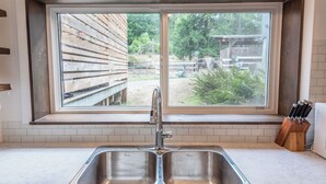 A kitchen with a double stainless steel sink, a window with a view of a wooden deck and greenery, and a knife block on the countertop.