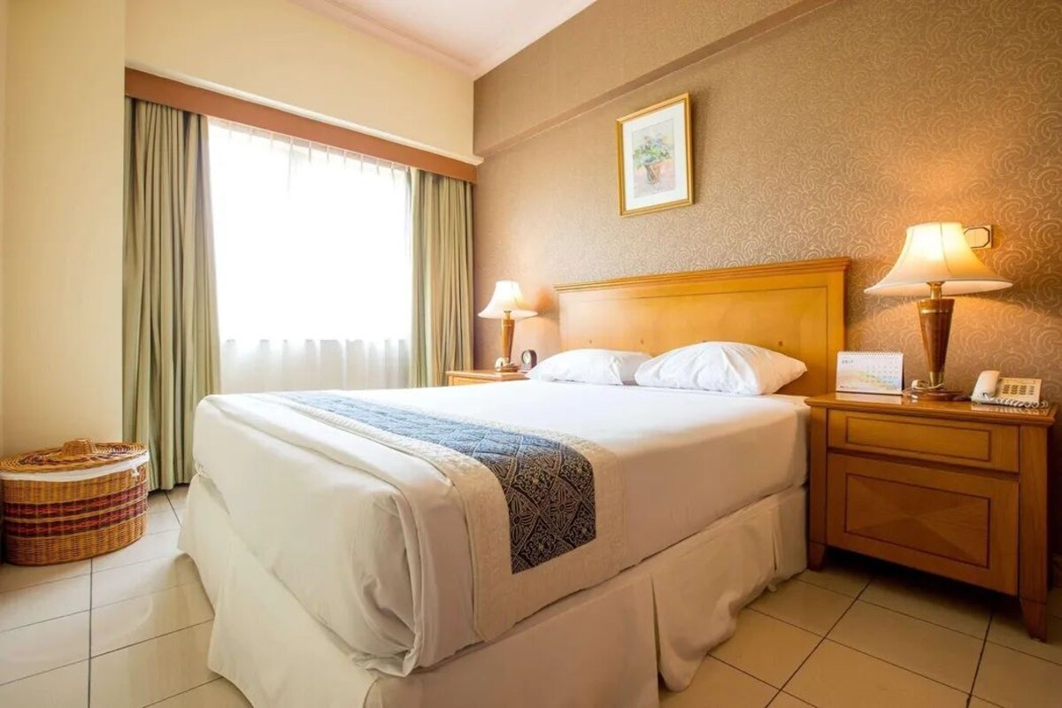 Habitación de hotel con una cama grande, dos mesitas de noche con lámparas, una mesita pequeña con un teléfono, una ventana con cortinas y un cuadro colgado en la pared.