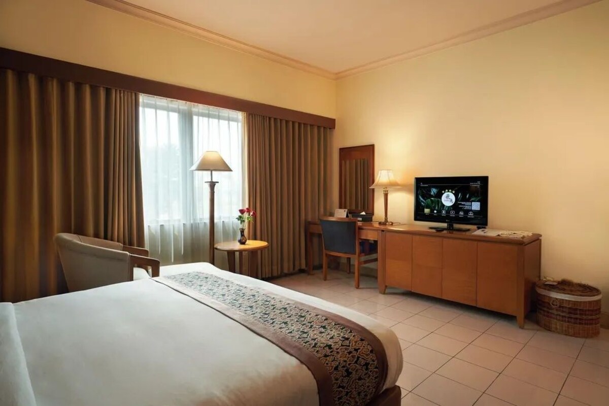 Habitación de hotel con cama, escritorio, silla, televisión y ventana con cortinas.