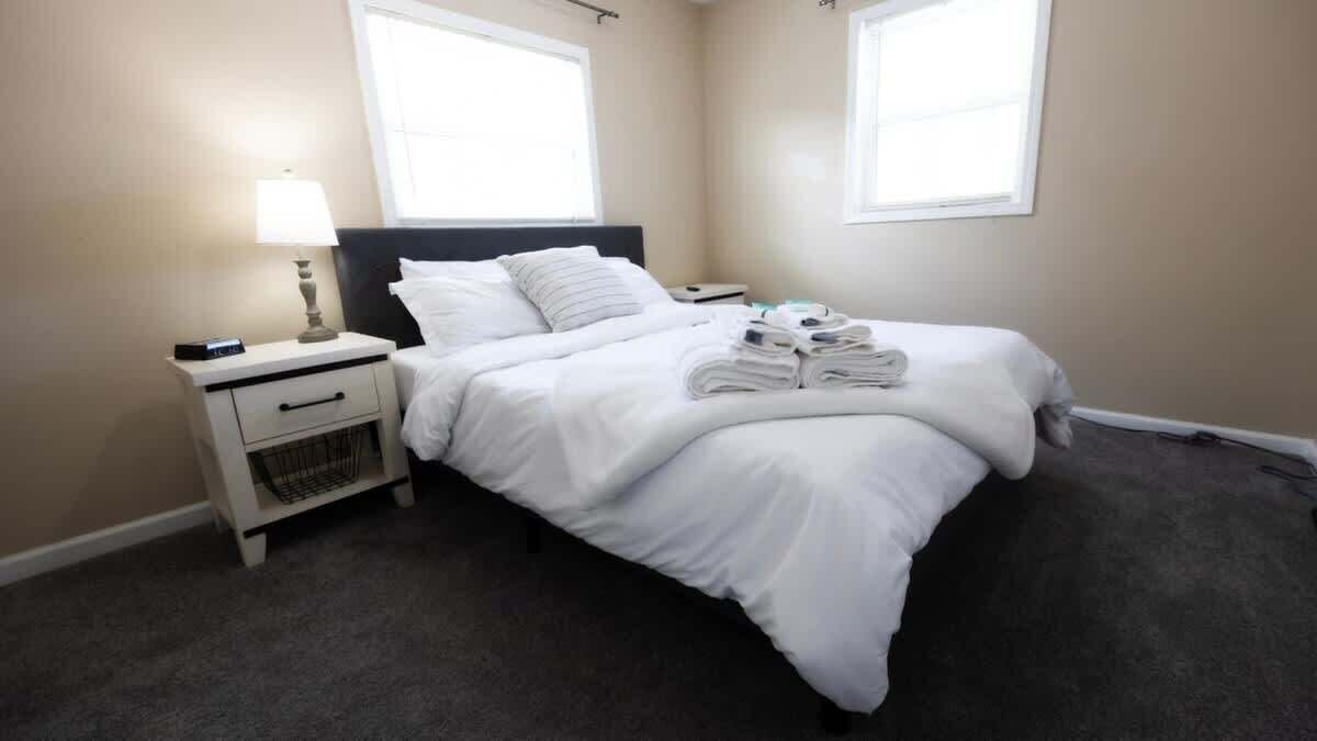 A neatly made bed with white linens, a bedside table with a lamp, and a small basket.