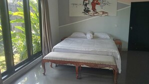 A bedroom with a large bed, a bedside table, a painting on the wall, and a window with curtains. - Villa Mertanadi 18 (Seminyak)
