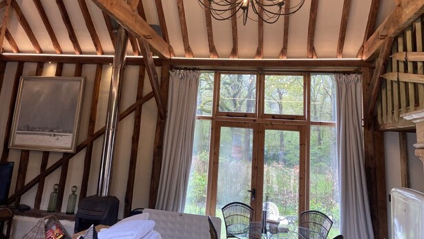 A rustic room with exposed wooden beams, a chandelier, a television, and a large window with curtains. - Beautiful one Bed Barn in national park 1 hr from London with small pool (Petworth)